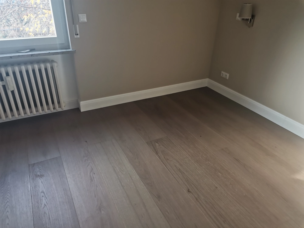 Leerer Raum mit frisch verlegten Holzdielen, weißen Sockelleisten, Heizkörper unter dem Fenster und Wandlampe – Bodenverlegung