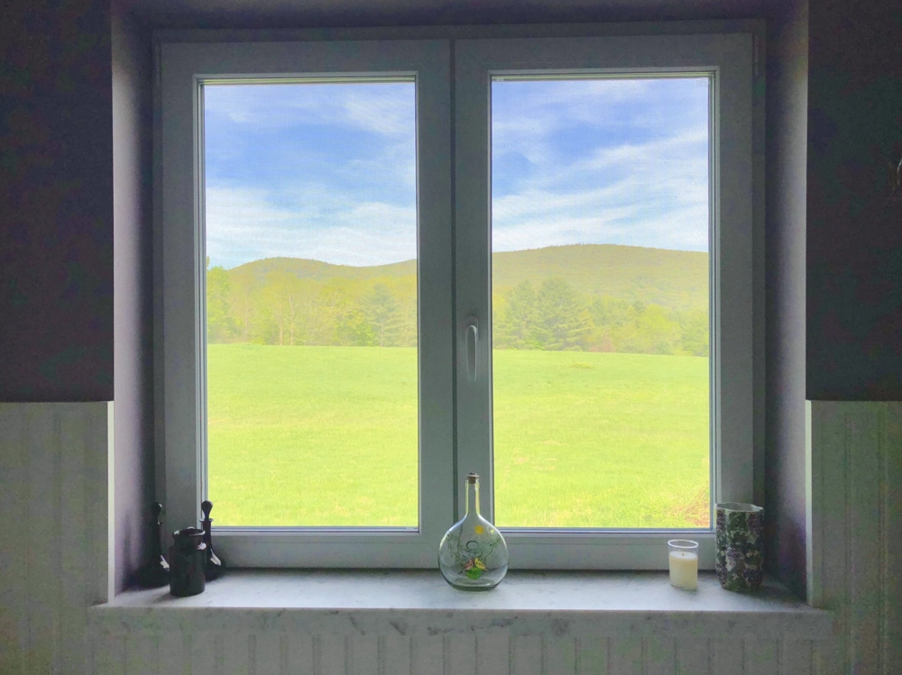 Weißes, doppelflügeliges Kunststofffenster mit Fensterbank und Dekoelementen vor grüner Wiesen- und Hügelkulisse – Fenstermontage