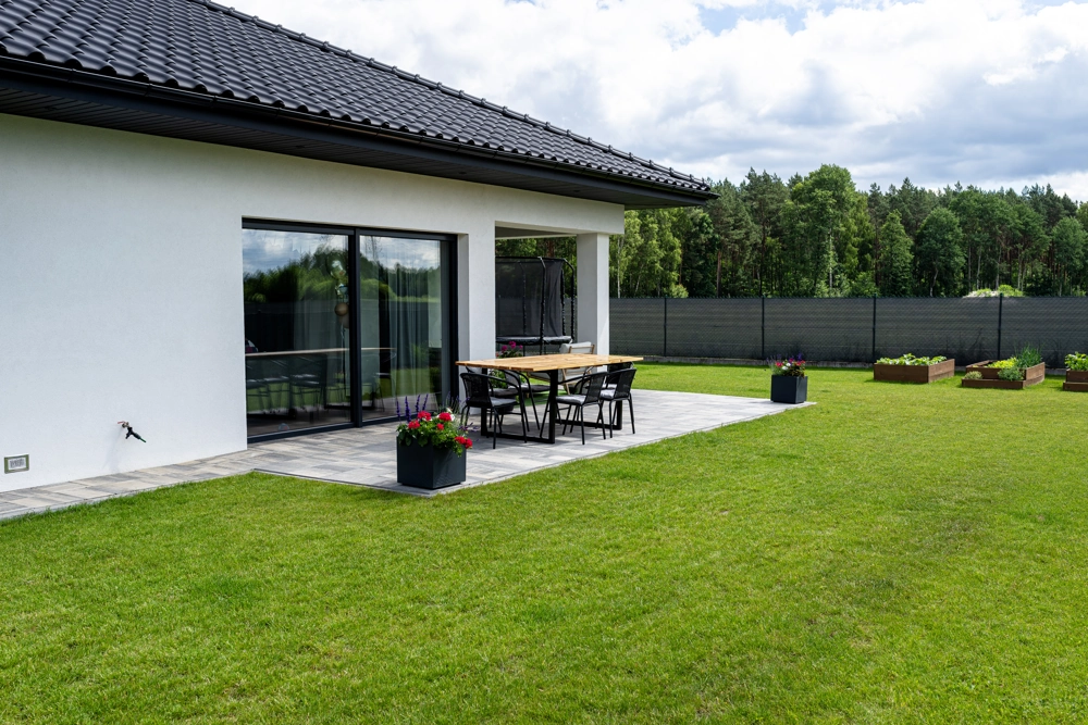 Gepflasterte Terrasse mit Holztisch und Stühlen vor weißer Fassade und gepflegtem Rasen Terrasse pflastern