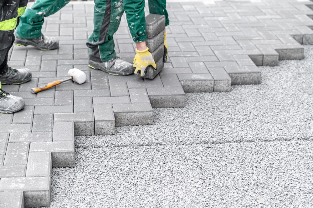 Zwei Handwerker verlegen graues Betonsteinpflaster auf Schotterbett, Gehwege pflastern