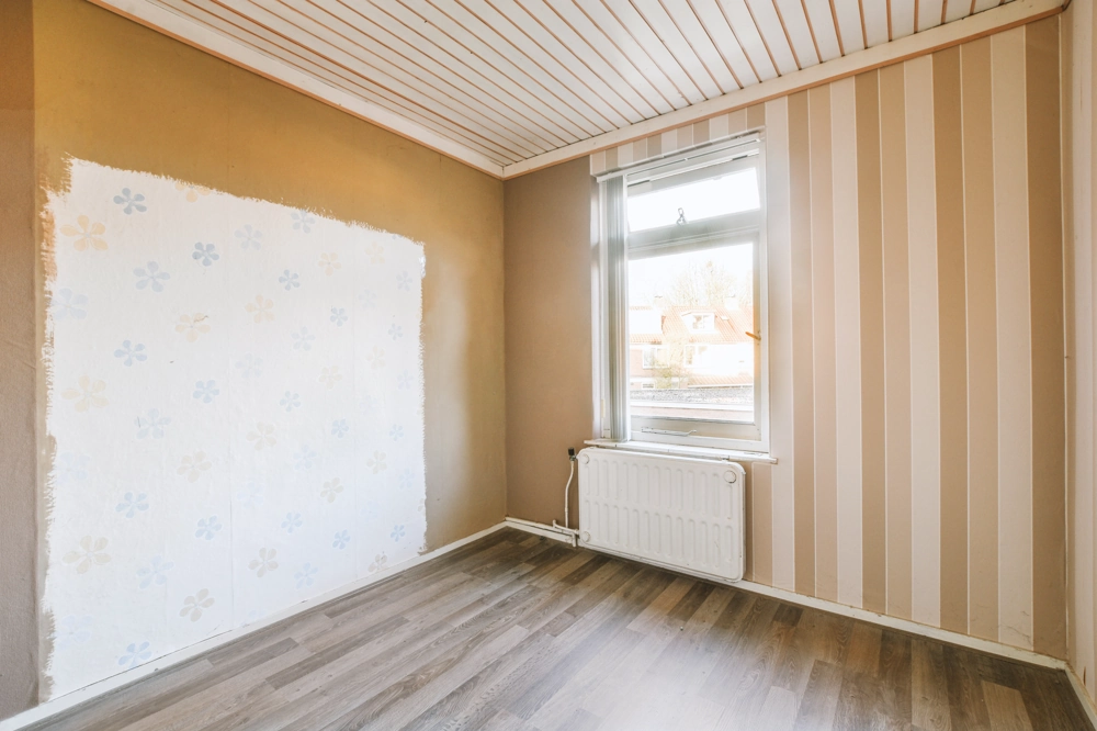 Leerer Raum mit Laminat-Bodenverlegung, teils freigelegten Wänden, Deckenpaneelen und Heizkörper unter Fenster