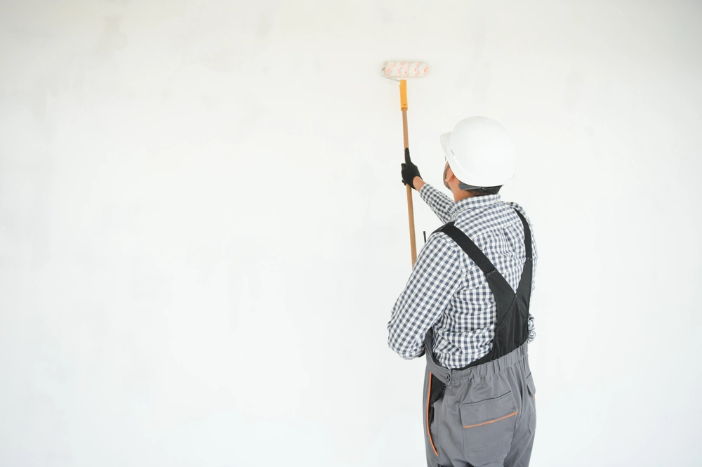 Handwerker in Schutzhelm und Overalls streicht mit Farbroller eine weiße Innenwand (Malerarbeiten)