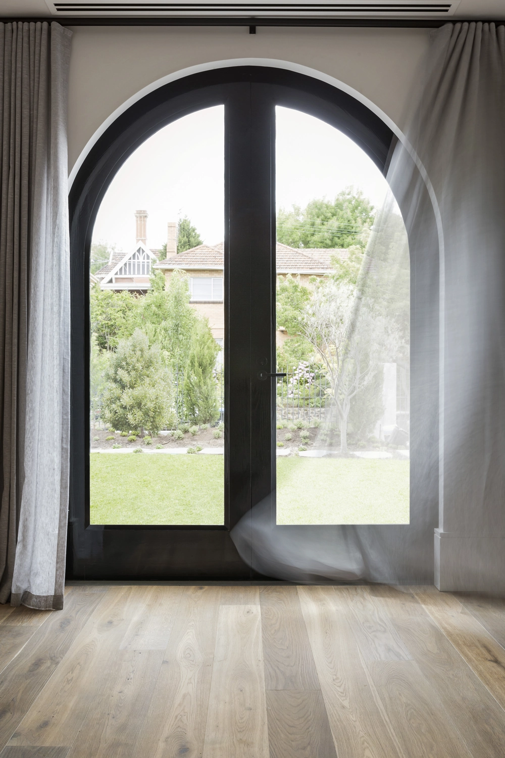 Schwarze, bogenförmige Glastür mit zarten Vorhängen und Blick auf Rasen und Garten, Fenstermontage