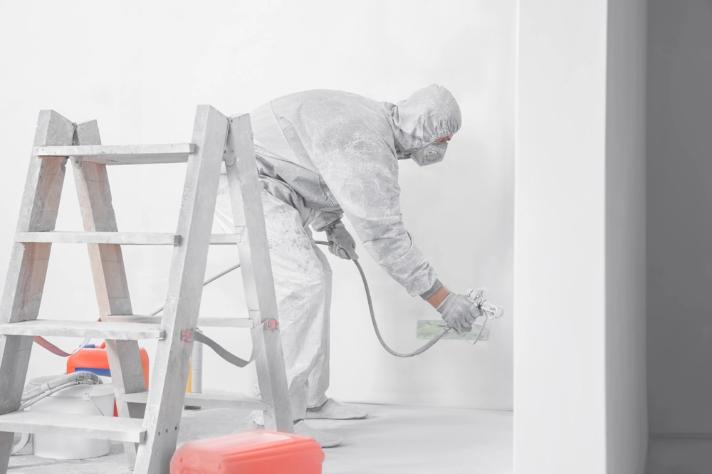 Handwerker in Schutzanzug sprüht weiße Farbe per Airless-Spritzgerät an Innenwand (Spritzlackierung)